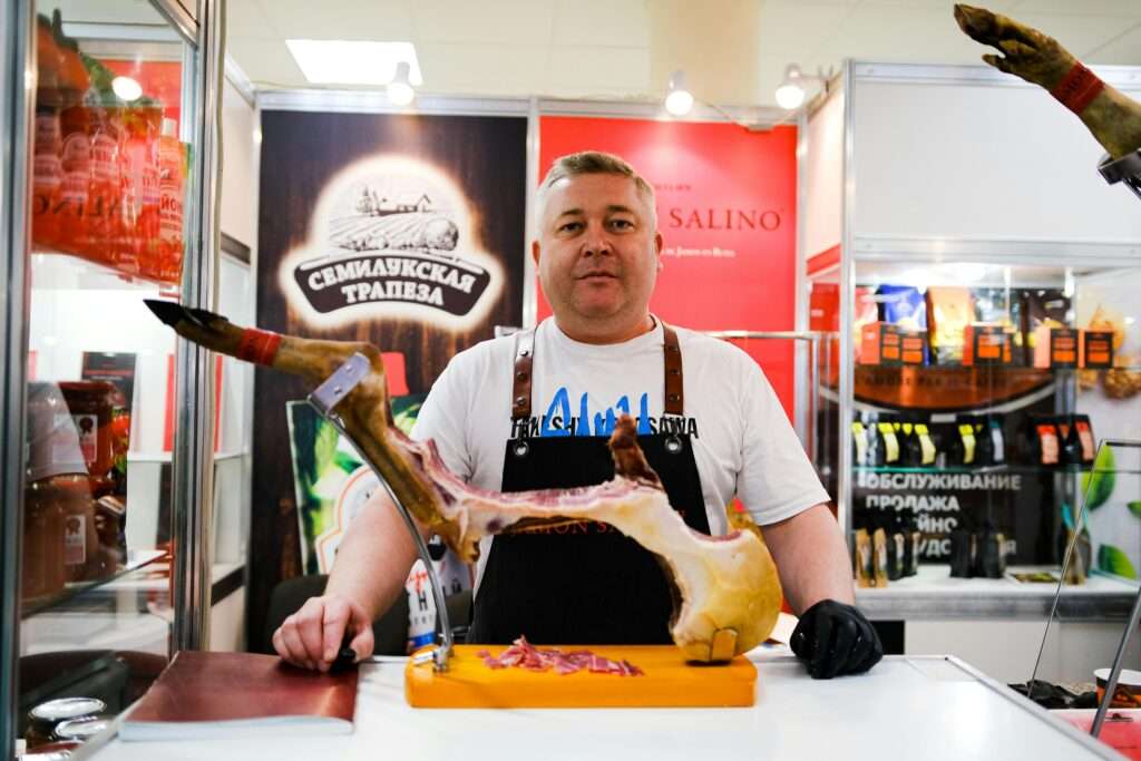 A butcher showcasing cured meat at an indoor trade show, promoting gourmet food products.