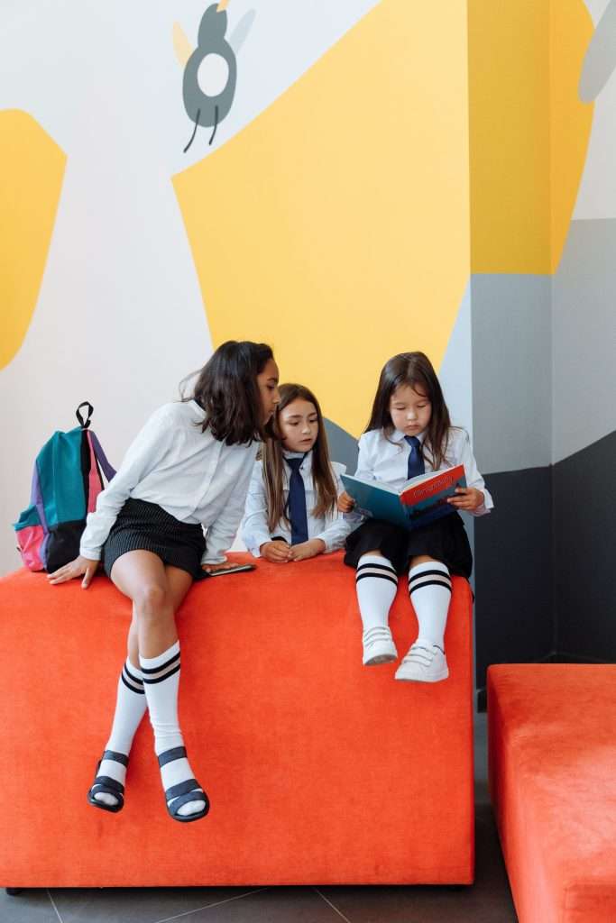 Three girls in school uniforms reading a book indoors, engaging in education.